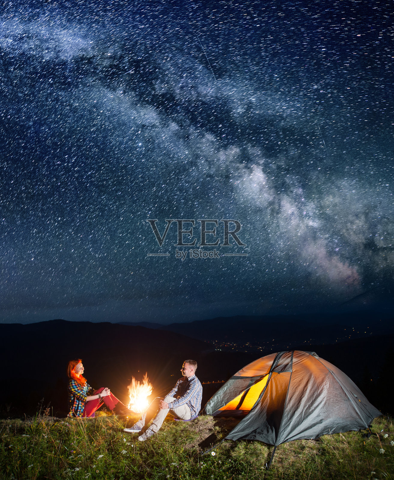 晚上露营。幸福的背包客夫妇坐在篝火和帐篷下，在难以置信的美丽星空下。村庄在山谷的背景。长时间曝光照片摄影图片