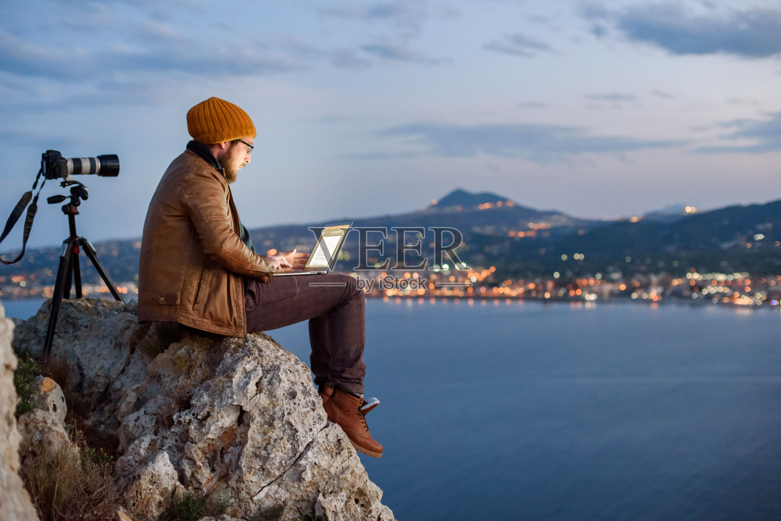 年轻迷人的摄影师坐在岩石上，用笔记本电脑拍摄大海和山脉的景色照片摄影图片