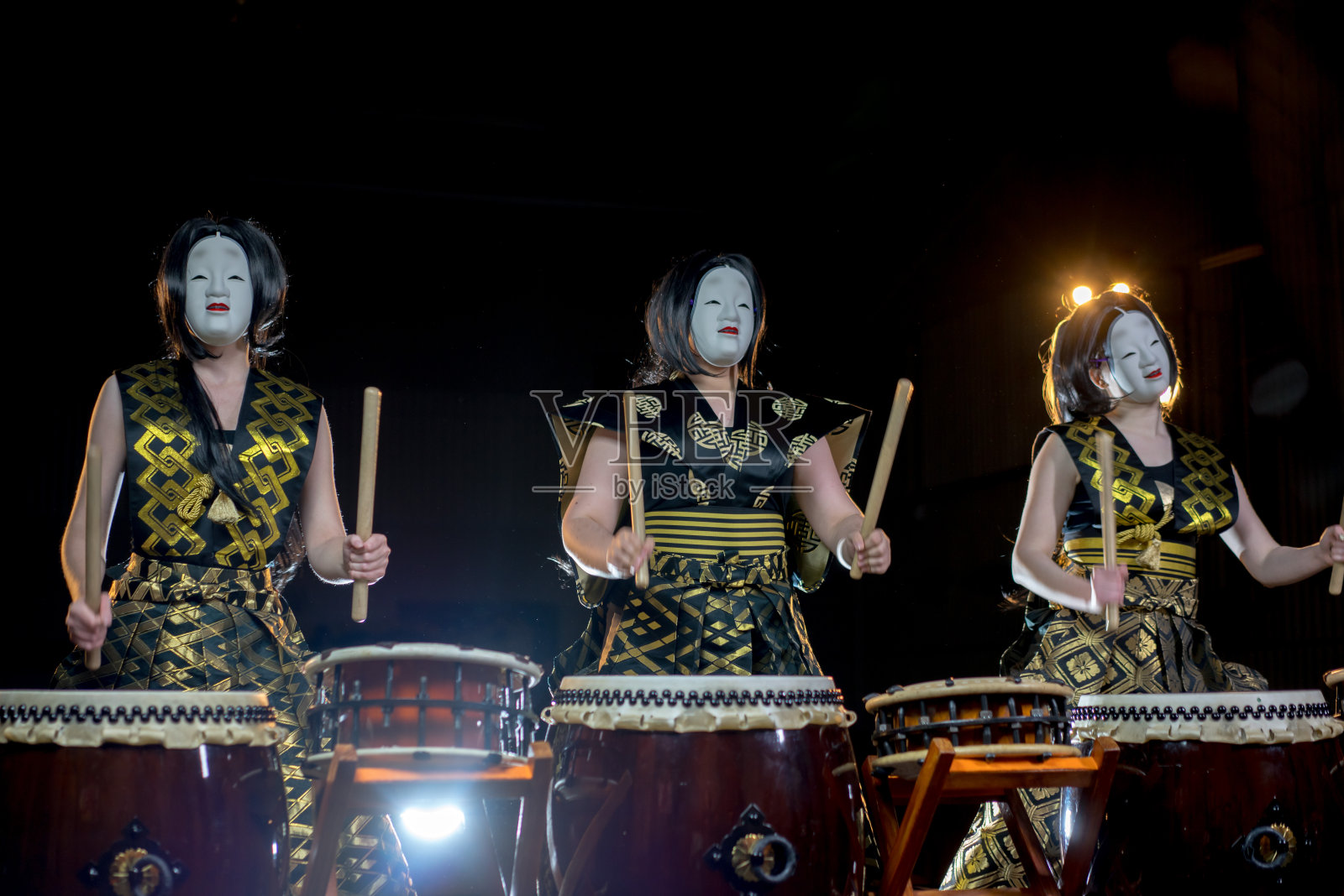 三个鼓手女孩戴着白色的恶魔面具和鼓槌，在黑暗的背景下拍摄。照片摄影图片