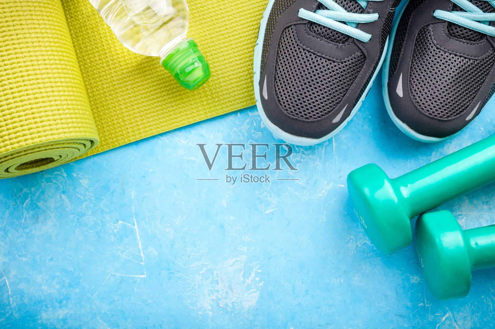 蓝色背景上的瑜伽垫、运动鞋、哑铃和一瓶水。健康生活方式、运动和饮食的概念。运动设备照片摄影图片