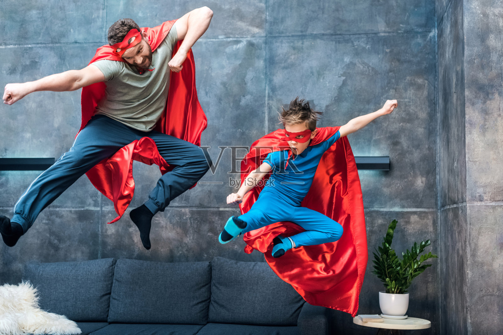 父亲和儿子穿着超级英雄服装在家里的沙发上跳照片摄影图片