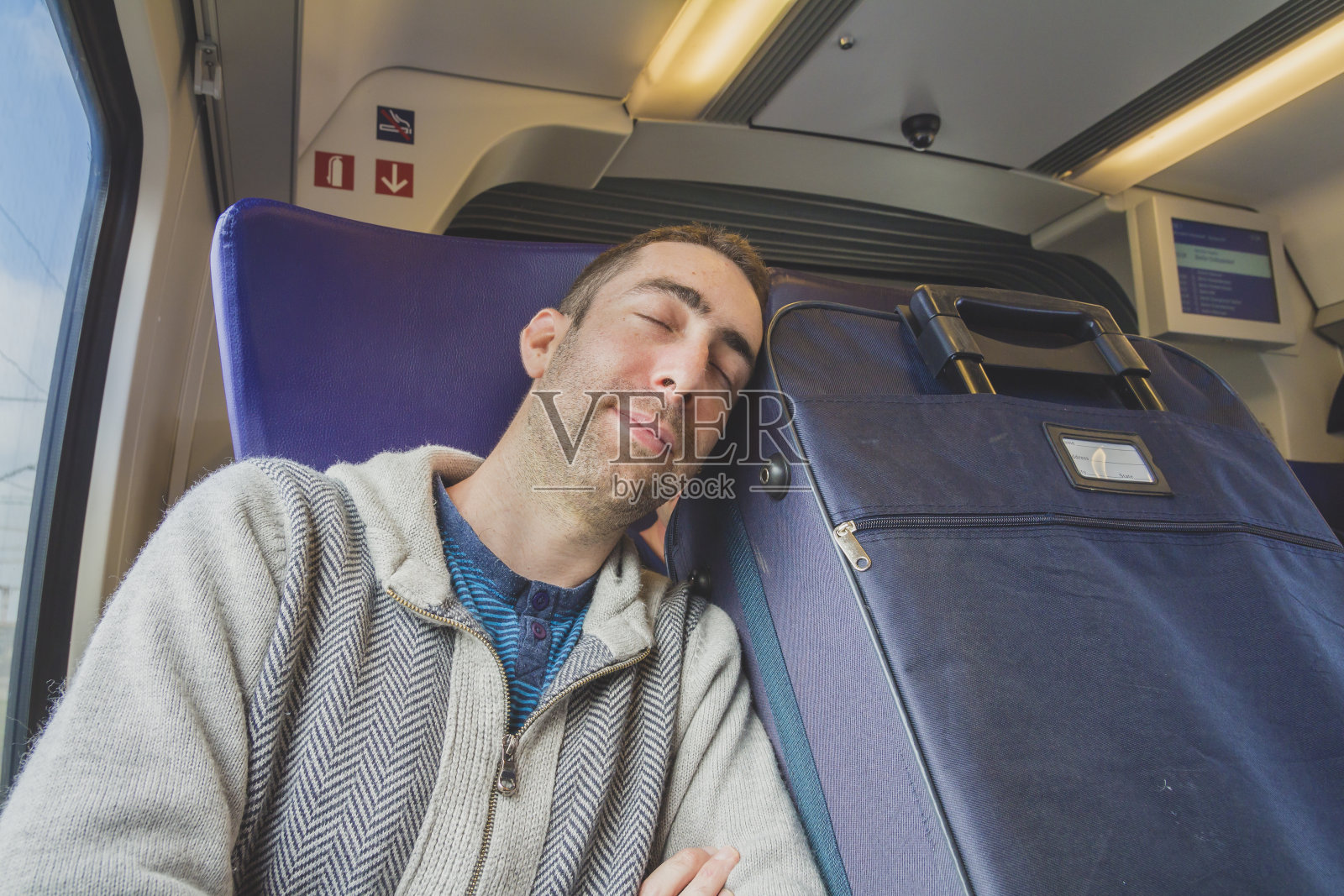 一个年轻人在火车上旅行，睡在他旁边的蓝色行李箱上照片摄影图片