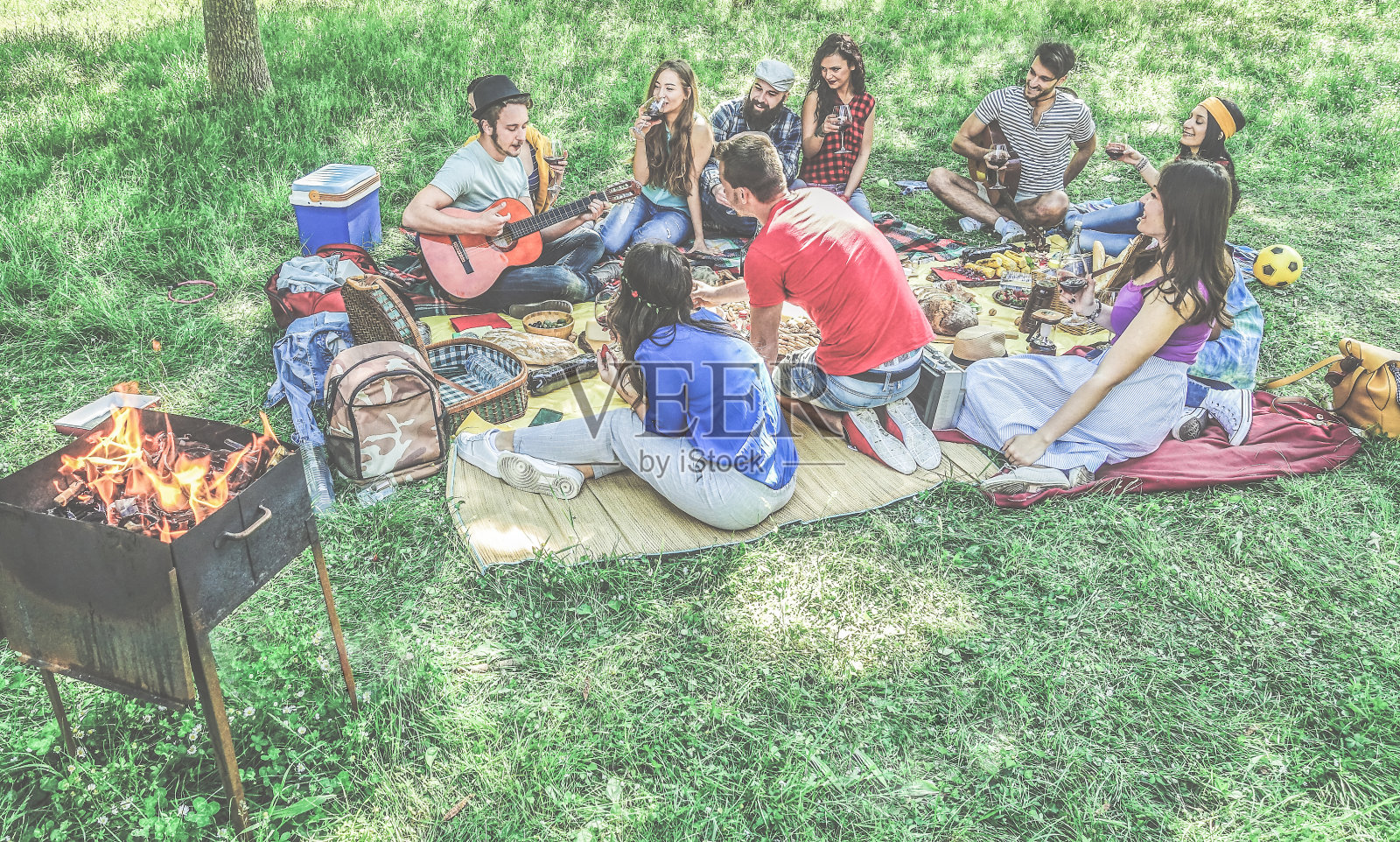 一群朋友野餐烧烤户外在城市公园-年轻人有乐趣玩音乐和放松的烧烤派对坐在草地上-主要集中在底部的家伙-对比过滤器照片摄影图片