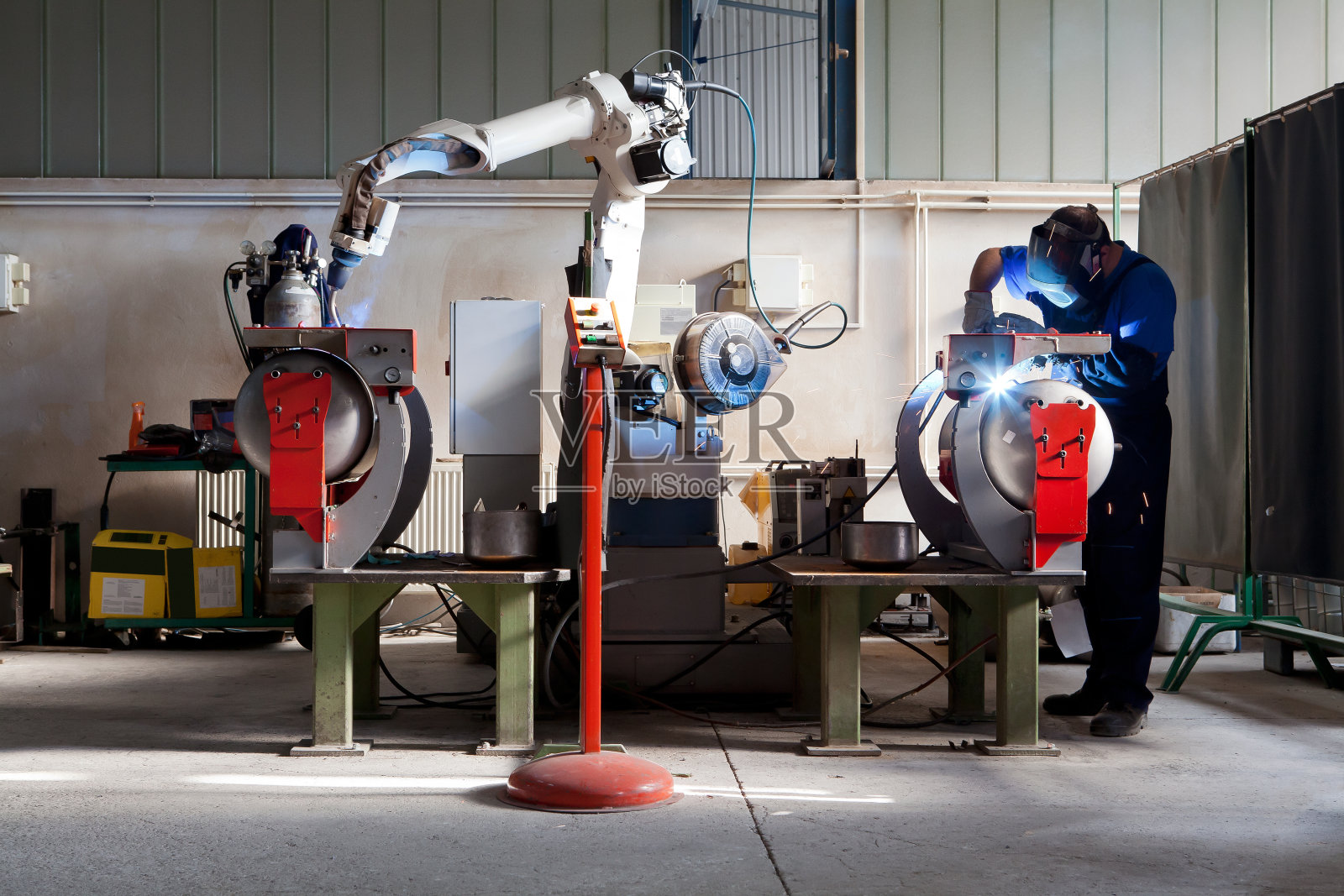 人与机器人在工业建筑中共同工作。机械臂在一名工人的协助下对金属部件进行焊接，该工人反过来手动管理焊接。照片摄影图片