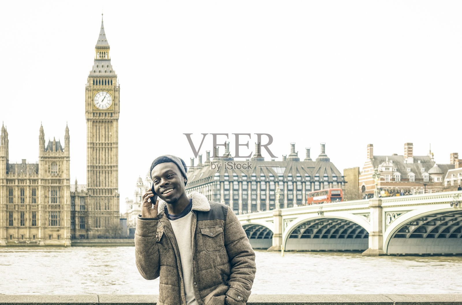 非裔美国年轻人在伦敦泰晤士河边使用智能手机-潮男男模穿着时髦的时尚服装拿着现代手机-明亮的去饱和滤镜照片摄影图片
