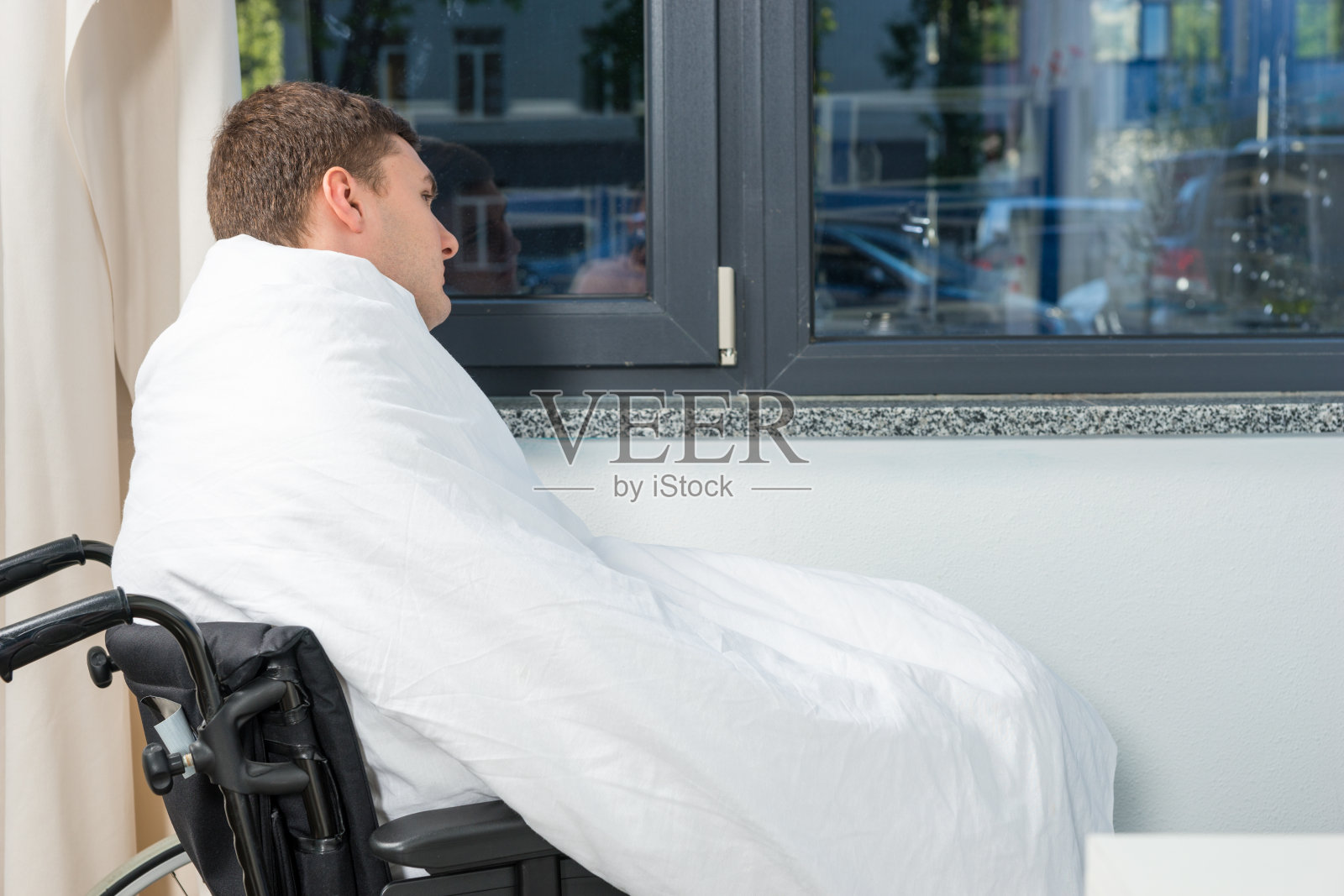 男病人坐在靠窗的轮椅上，盖着被子躺在病房里照片摄影图片