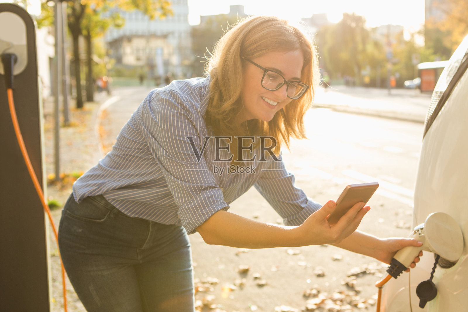 年轻女子站在电动汽车附近，手里拿着智能手机照片摄影图片