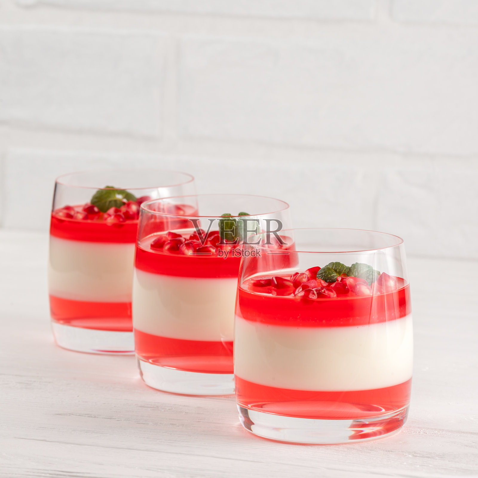 奶油香草意式奶冻配红色果冻在美丽的玻璃杯里，新鲜成熟的石榴在白色的木质背景上。照片摄影图片