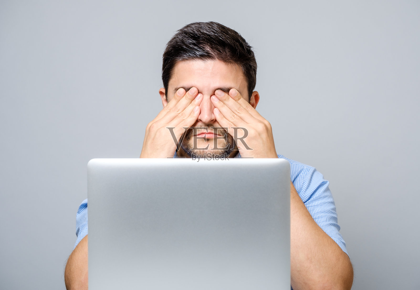 英俊的年轻人与笔记本电脑摩擦他的眼睛在灰色的背景照片摄影图片