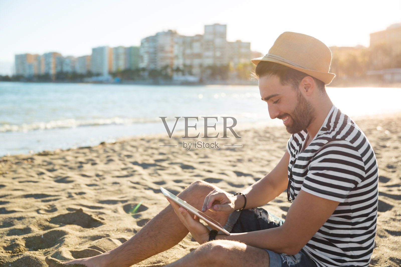 戴着帽子坐在沙滩上用平板电脑的快乐男人照片摄影图片