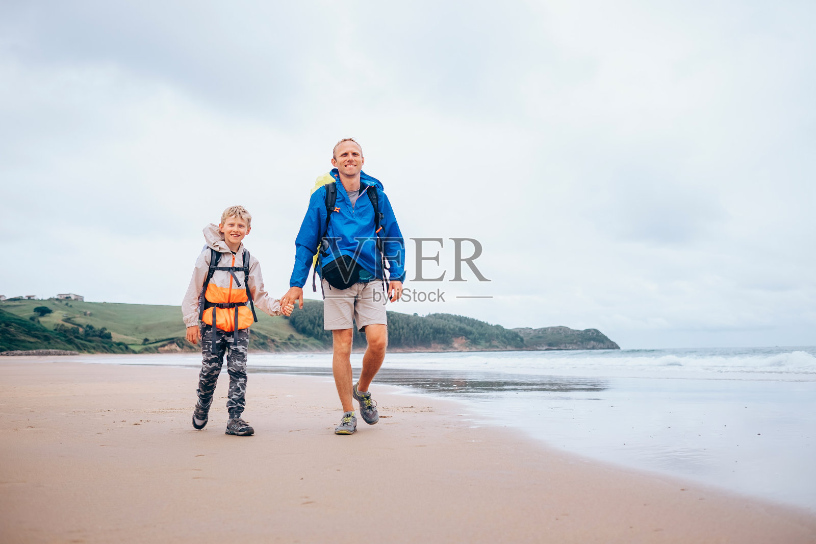 带着孩子旅行:父亲和儿子背着背包走在沙滩上，下雨天照片摄影图片