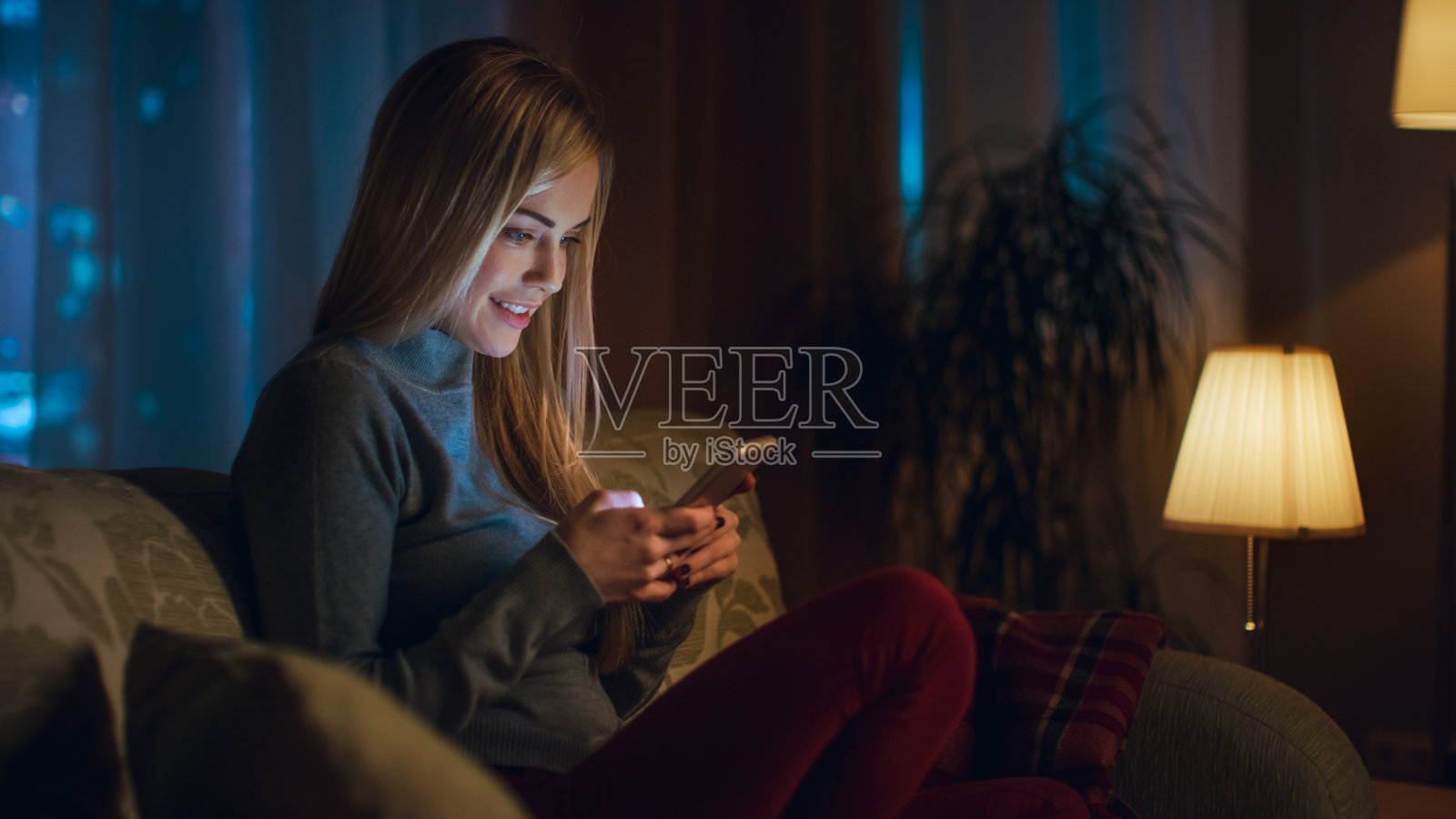 美丽的年轻女子坐在她的沙发在客厅。她有兴趣使用智能手机。傍晚时分，窗外是大城市。照片摄影图片