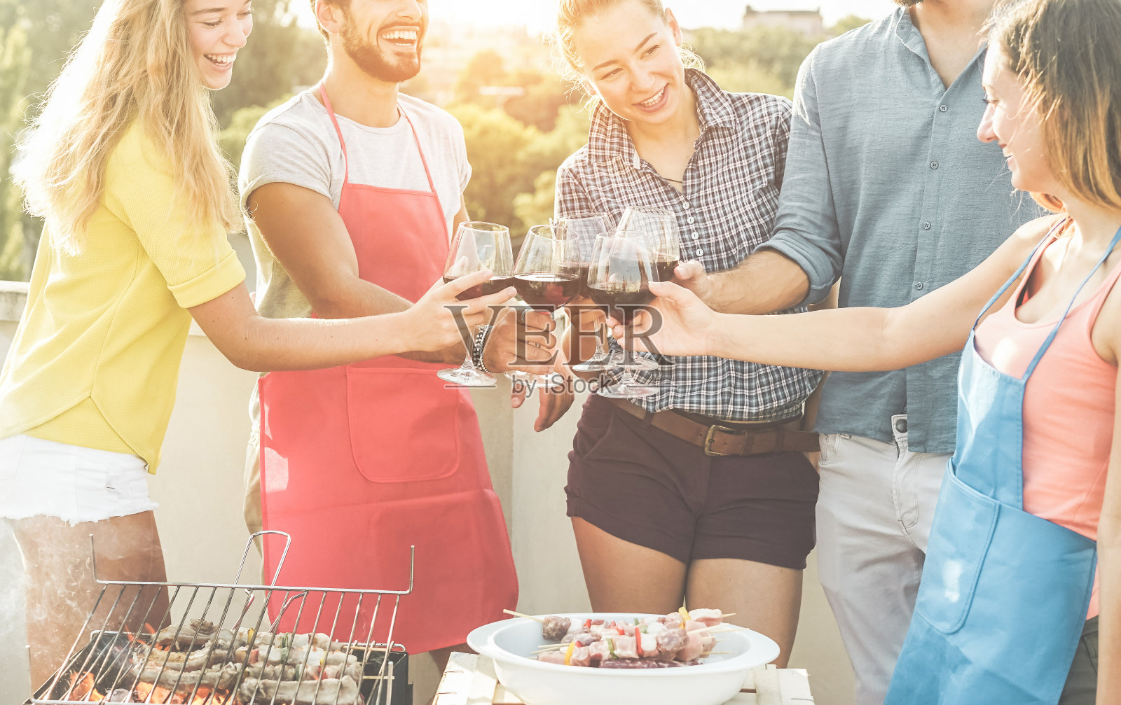 快乐的朋友们在烧烤晚宴上用红酒欢呼-年轻人在户外烧烤，夕阳为背景-聚焦眼镜-食物，乐趣，青年的生活方式和友谊的概念照片摄影图片