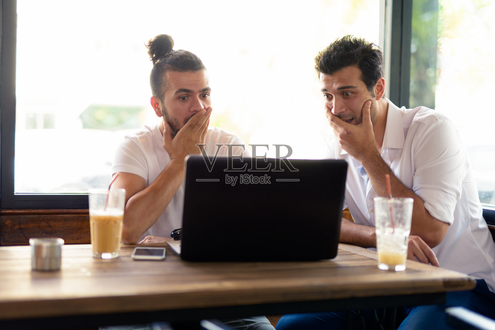 两名男子坐在咖啡店里，一边使用笔记本电脑，一边看起来很震惊，用手捂着嘴照片摄影图片