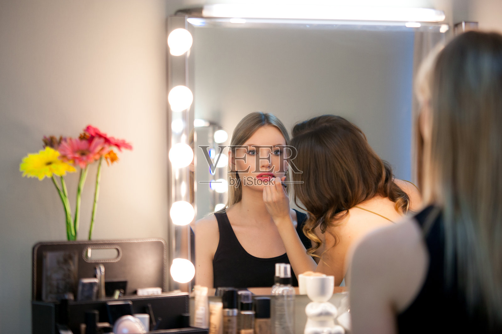 化妆师在美容工作室的镜子前用口红涂抹迷人的金发女孩。照片摄影图片
