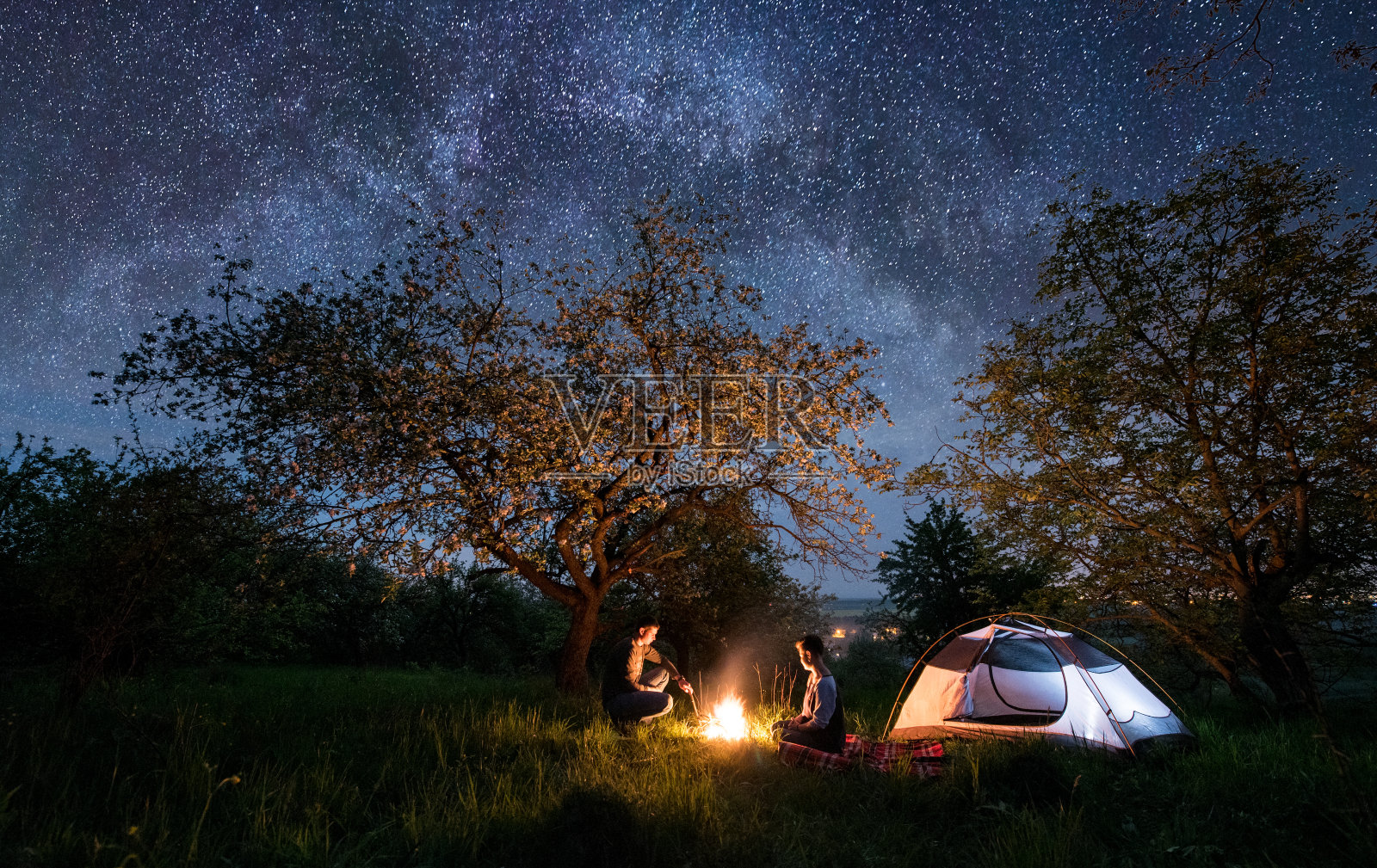 浪漫的情侣游客坐在篝火附近的帐篷下的树和美丽的夜空满是星星和银河。晚上露营照片摄影图片
