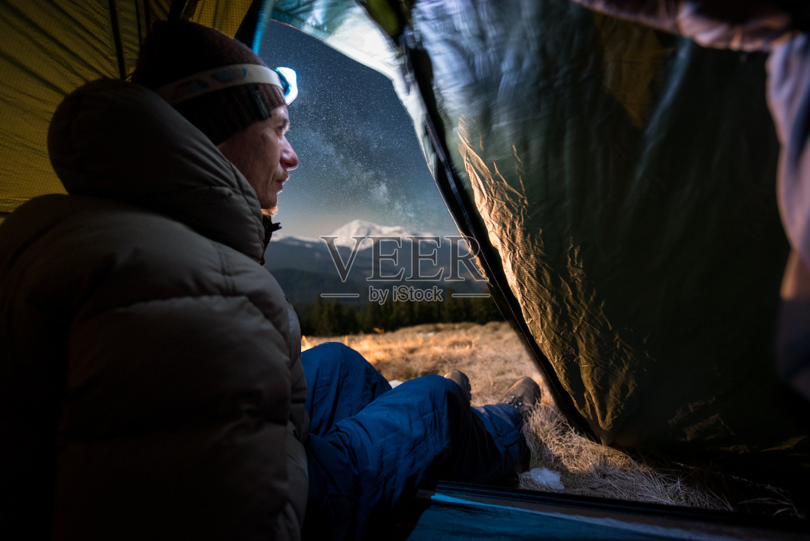 从帐篷里看，男游客在他的露营中享受夜晚。在满是星星和银河的美丽夜空下，一个带着头灯的男人坐在篝火旁的帐篷里照片摄影图片