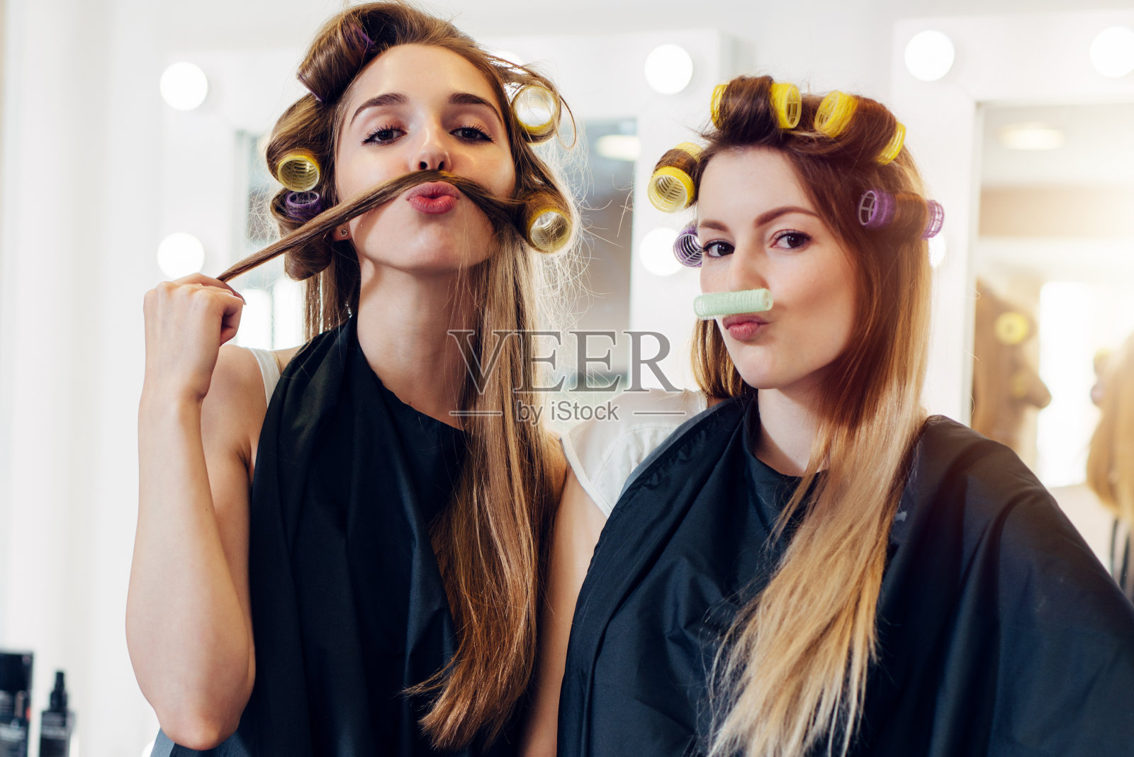 两个可爱的女朋友，穿着黑色的斗篷，戴着卷发器，在美发沙龙用她们的头发和卷发器做小胡子照片摄影图片