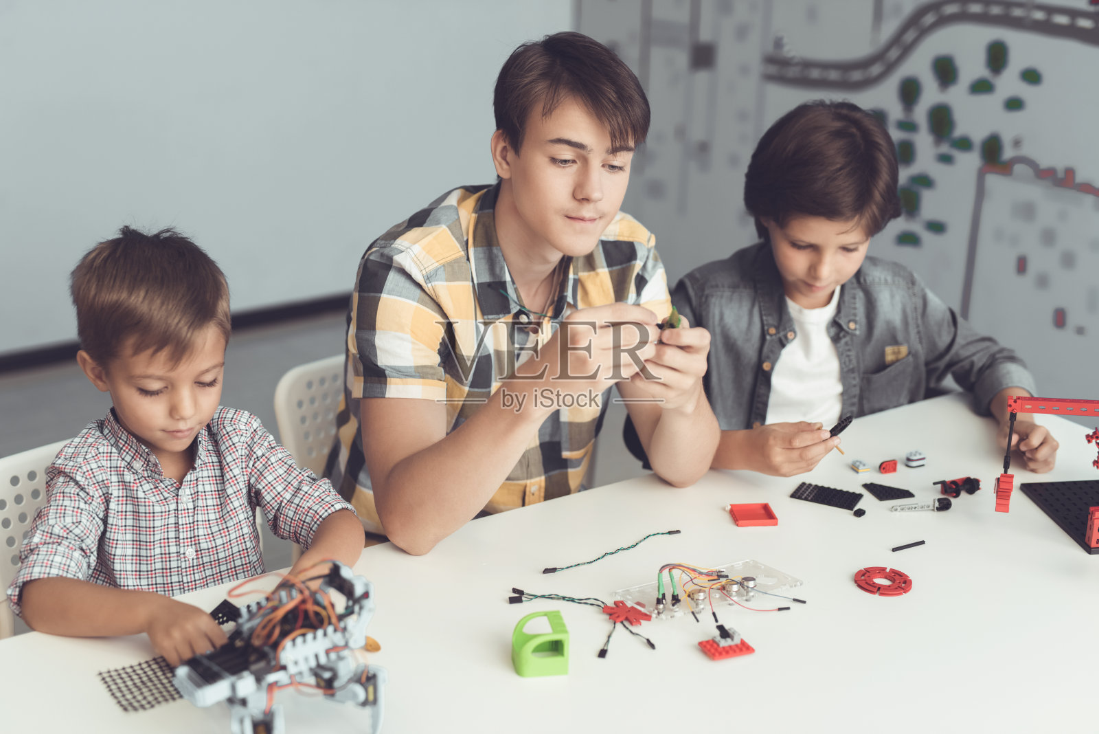 一个男人和两个男孩坐在一张灰色的大桌子旁建造一个机器人。在他们之前，有许多零件和组件照片摄影图片