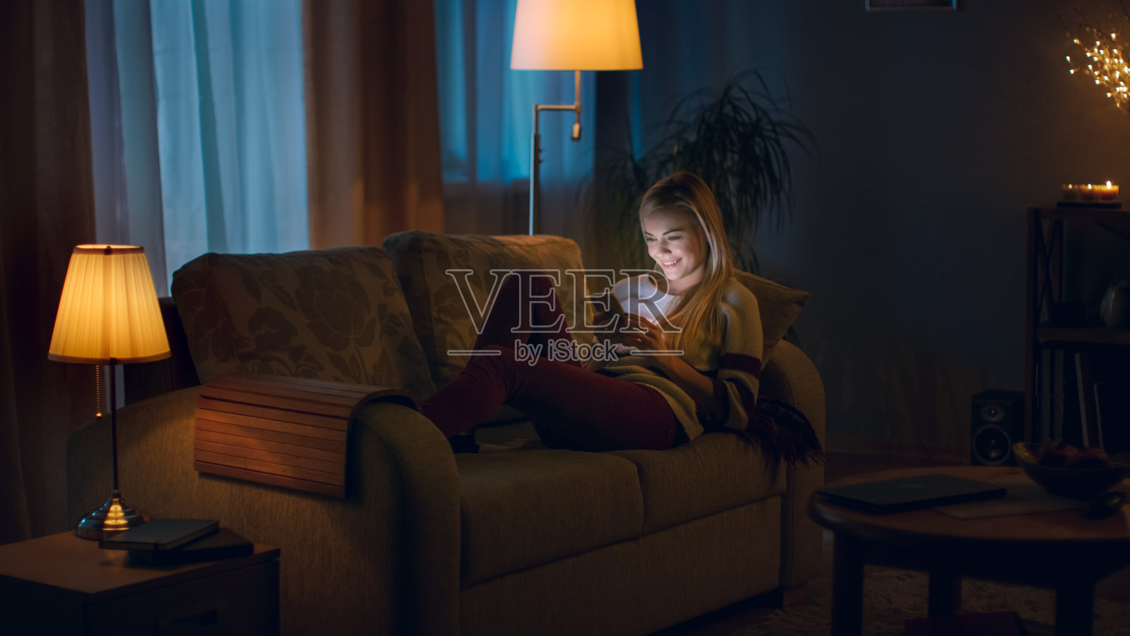 晚上，美丽的年轻女子躺在沙发上使用智能手机。房间看起来温暖舒适。照片摄影图片