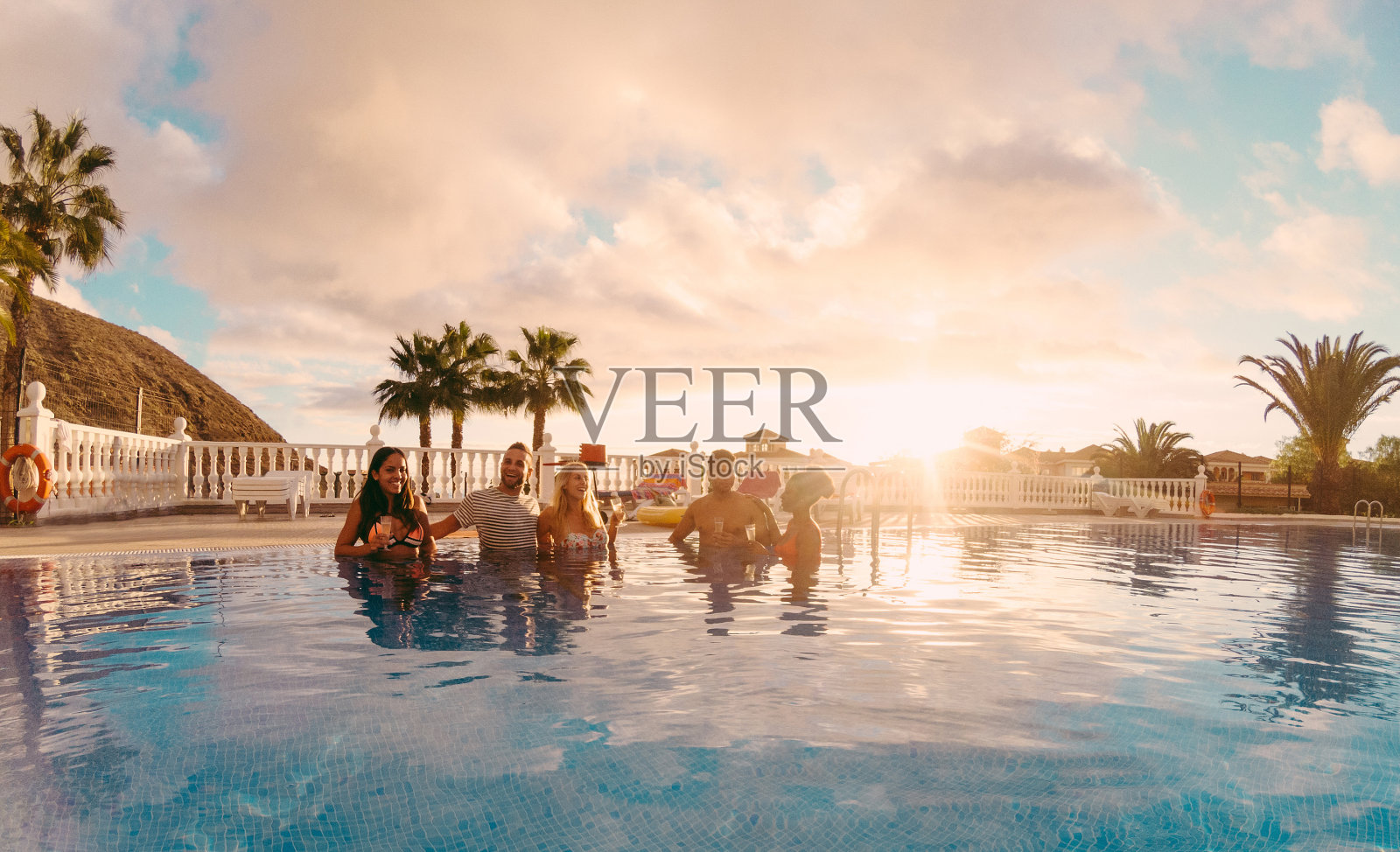 快乐的朋友喝香槟在游泳池聚会在日落-富有的人有乐趣在专属的热带度假-假日和友谊概念-主要集中在左边的家伙-原始的太阳颜色色调照片摄影图片