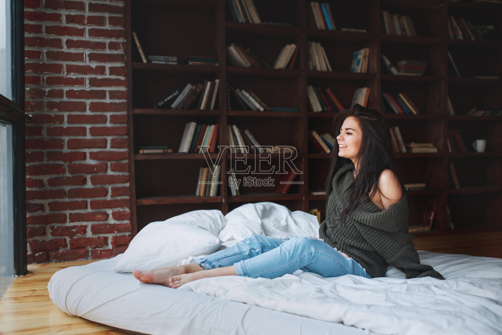 性感的年轻女子深色头发，绿色毛衣和牛仔裤坐在家里的床上照片摄影图片