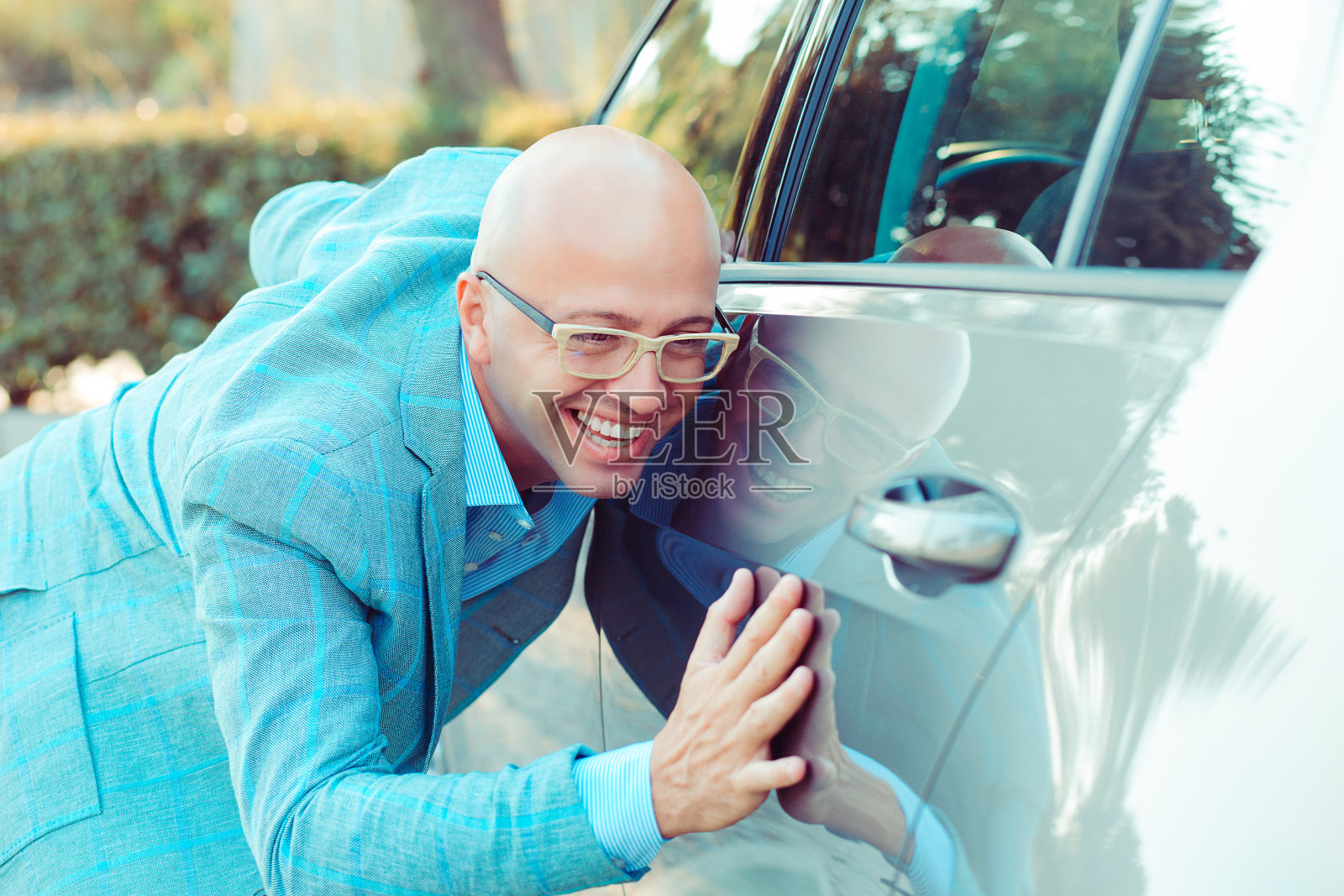 快乐兴奋的年轻秃头司机拥抱爱抚他的车在户外。照片摄影图片