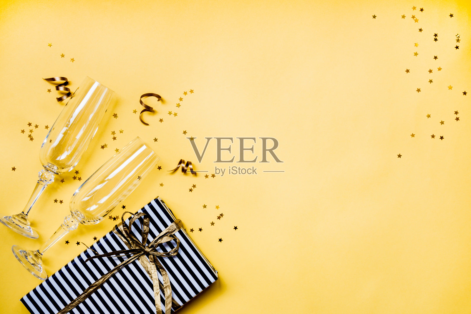 庆典背景-俯视图两个水晶香槟酒杯，一个礼盒包装在黑白条纹纸，丝带和星星形状的金色五彩纸屑在黄色的背景。副本的空间。照片摄影图片