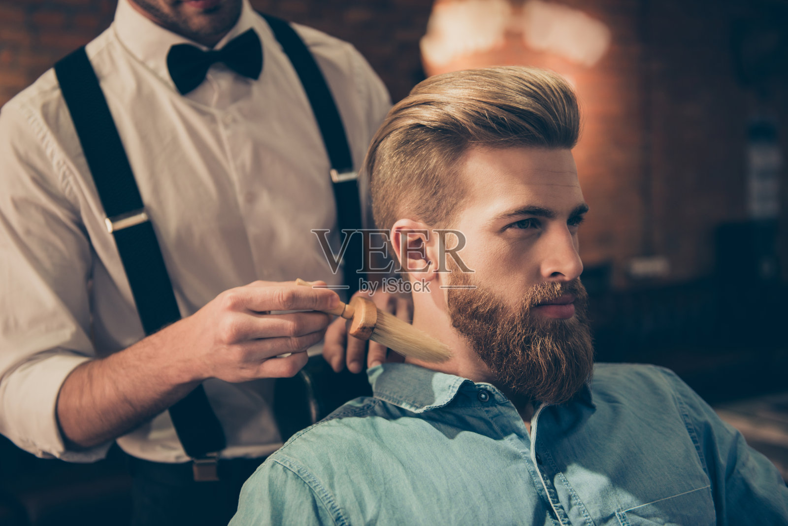 一名穿着考究的理发师正在用刷子清洁顾客的脖子，并为他展示自己的工作。惊人的!发型看起来很时尚，很完美照片摄影图片
