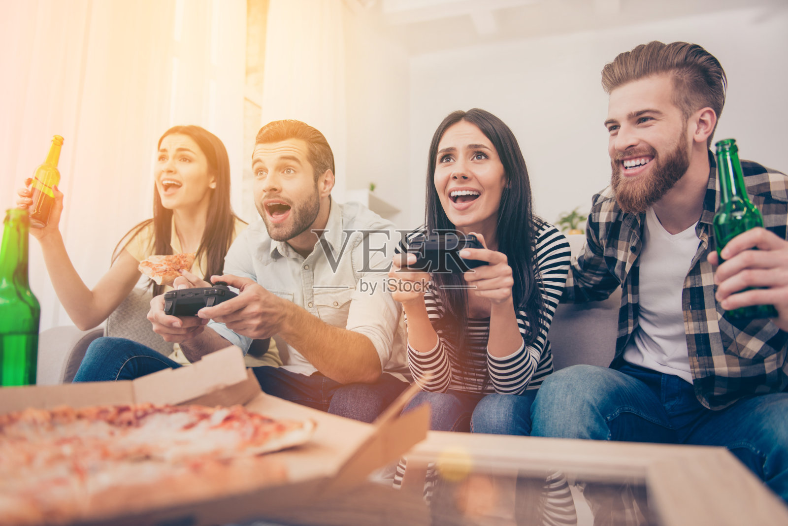 兴奋的朋友们坐在家里的沙发上玩电子游戏，喝着啤酒吃着披萨。他们在室内有很好的同伴度过了美好的周末照片摄影图片