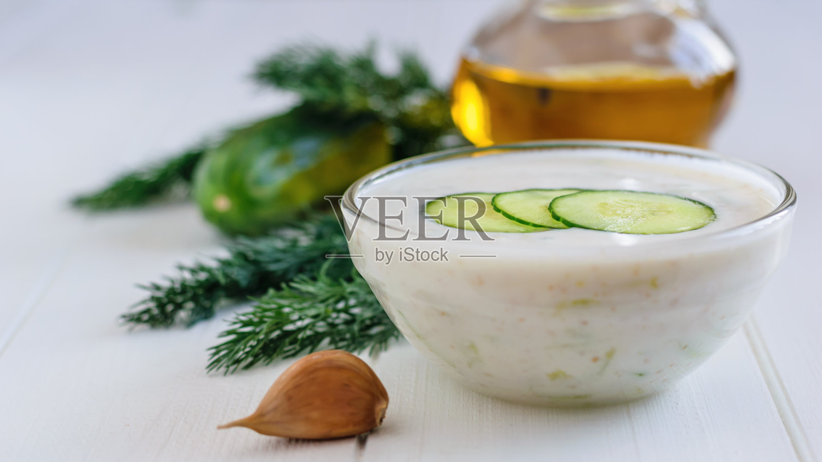 新鲜制作的酸奶黄瓜放在一个玻璃碗里，配上大蒜和欧芹放在白色木桌上。照片摄影图片