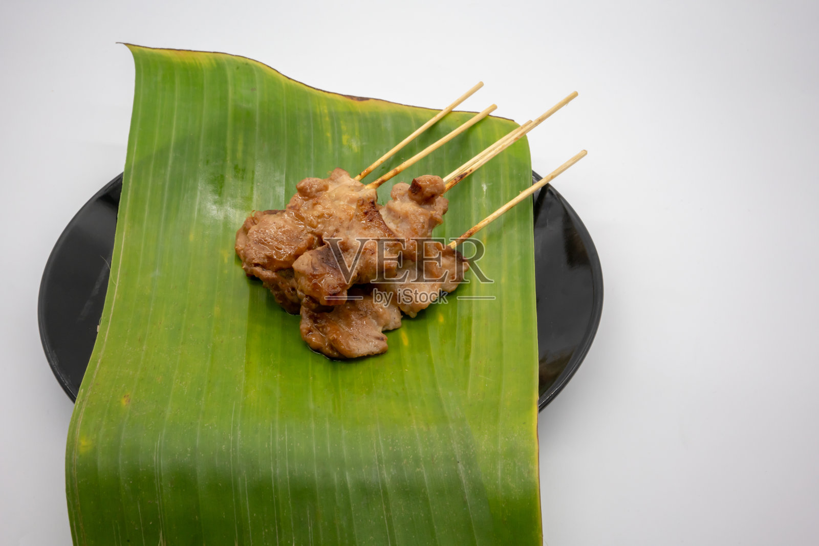 泰式烧烤或“Moo Ping”在泰国的单词。香蕉叶上用木棒塞的腌猪肉，白色背景上的黑色陶瓷盘。照片摄影图片