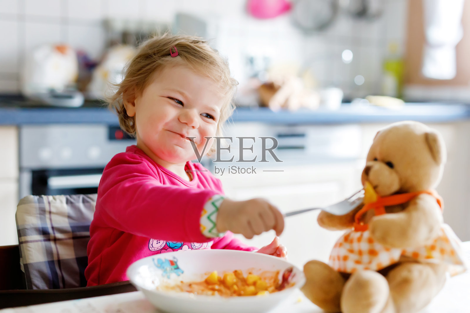 可爱的小女孩用叉子吃蔬菜和意大利面。小孩子在喂玩具熊，玩玩具熊。照片摄影图片