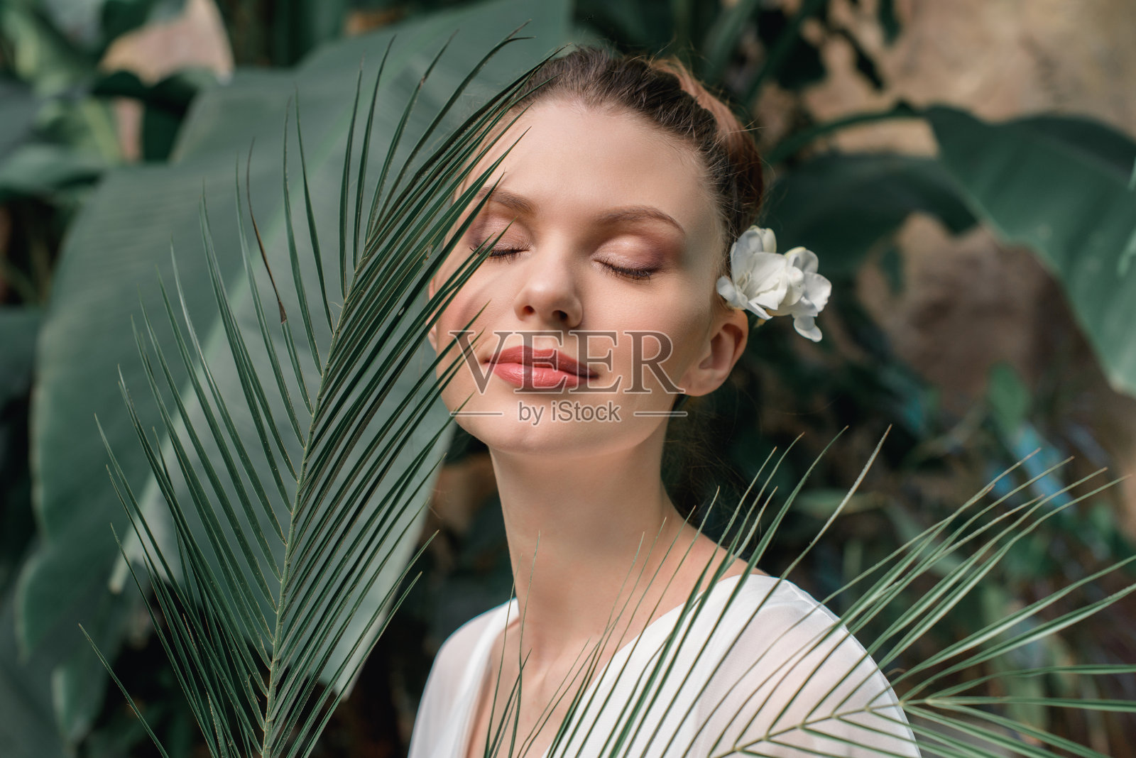 美丽的女孩闭着眼睛摆姿势，白色的夏装与热带棕榈叶照片摄影图片