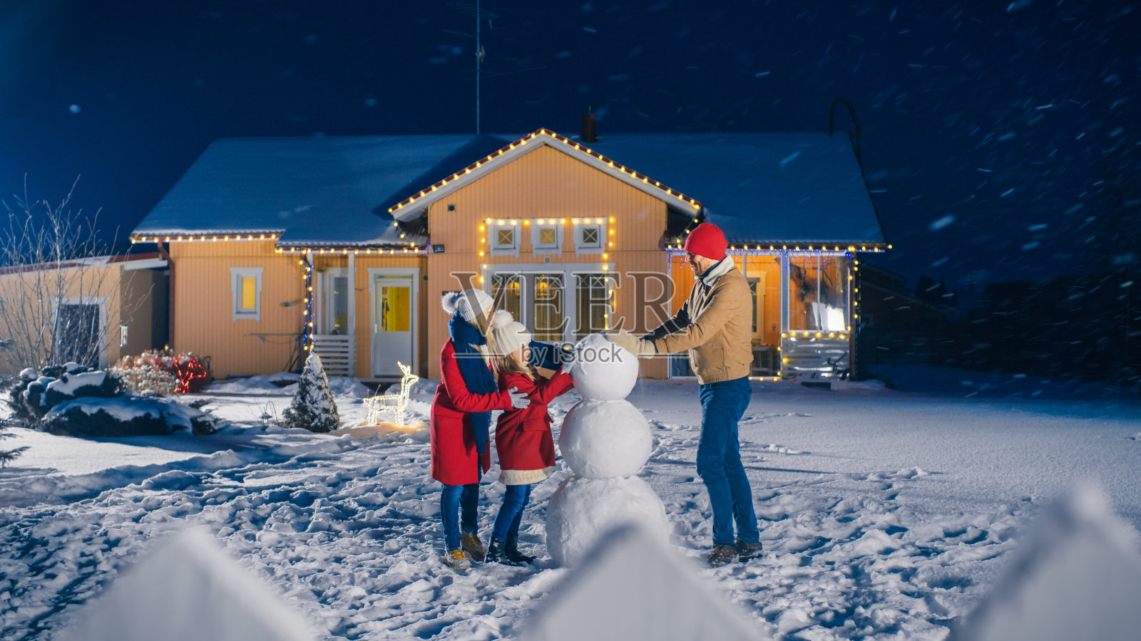 快乐的年轻家庭在他们田园般的房子的后院堆雪人。爸爸让雪球、女儿和妻子帮助他。一个冬天的晚上，家人在一起。照片摄影图片