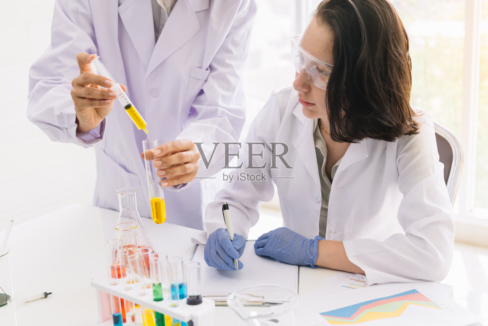 实验室里的年轻女科学家正在记录测试结果，显示一个黄色荧光试管和同事们在滴液体。照片摄影图片