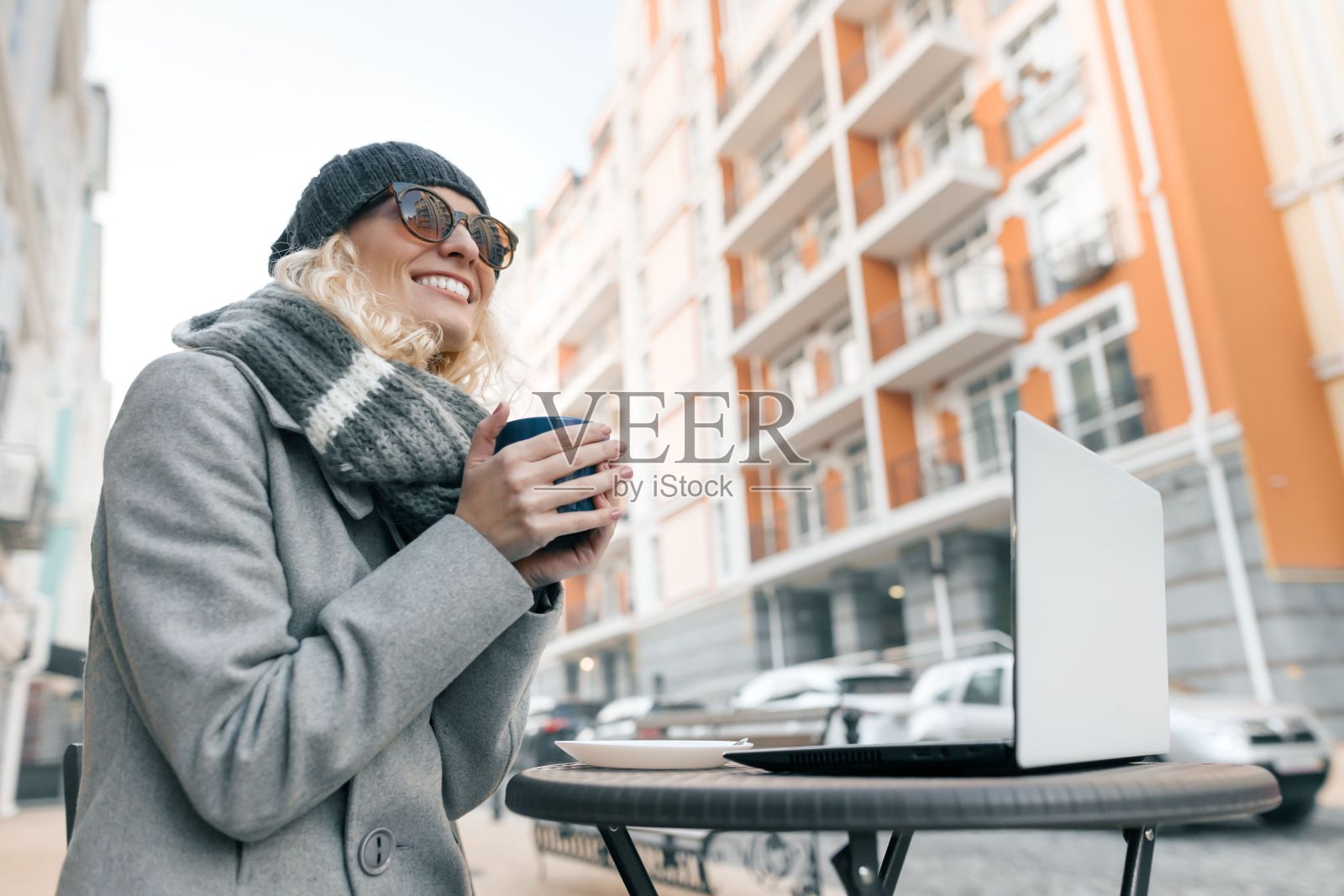 年轻的微笑妇女的肖像在温暖的衣服，编织的帽子。女孩暖手与一杯热饮料在一个户外咖啡馆在城市街道的背景照片摄影图片