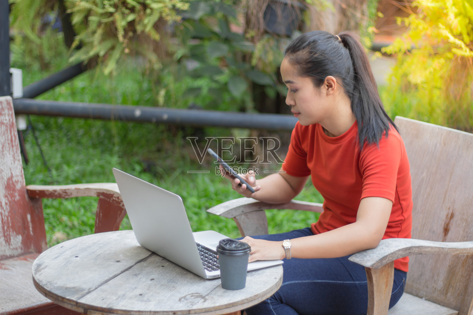 女人微笑着玩手机，笔记本电脑和咖啡杯放在木桌上。照片摄影图片