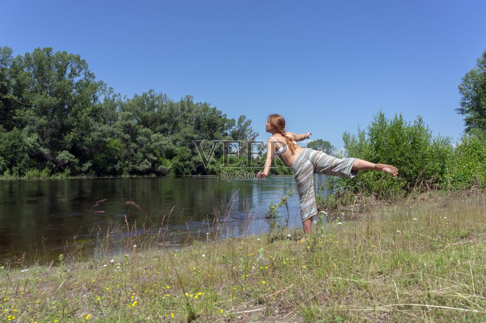 在一个阳光明媚的夏日，一个红头发的女孩在河边做体操，背景是树木。照片摄影图片