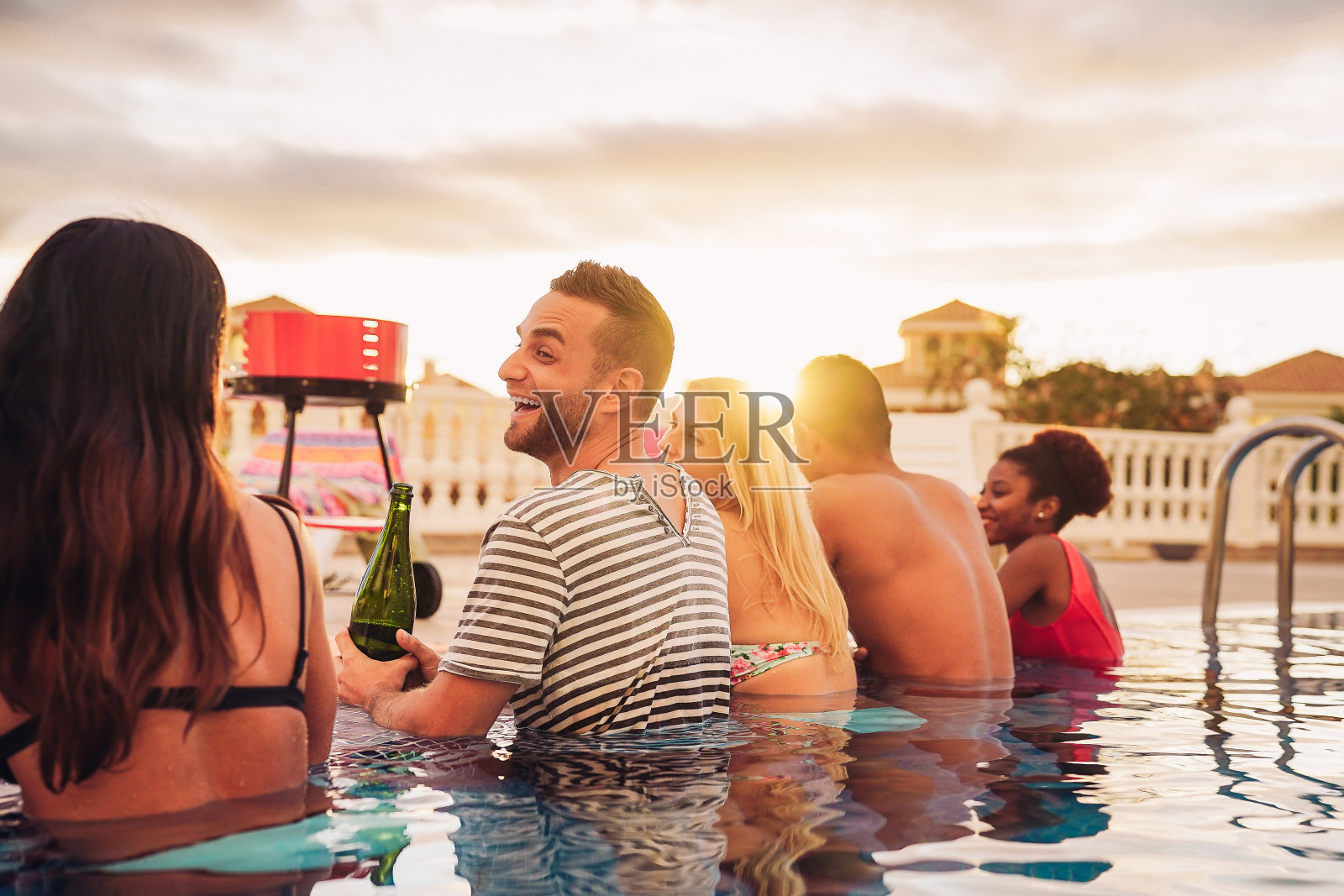 快乐的朋友使一个游泳池聚会喝香槟在泳池边-年轻人在日落的豪华热带度假村烧烤晚餐的乐趣-青年假日的生活方式概念照片摄影图片