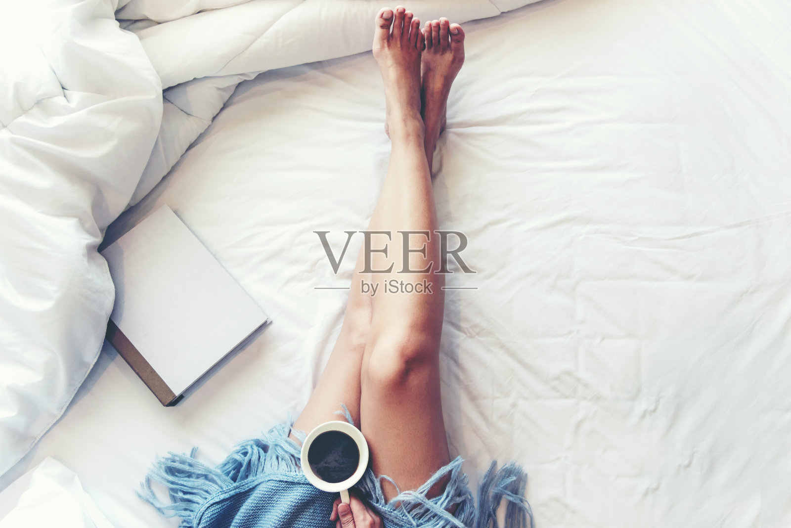 闭上双腿的女人在白色的床上。女人在早晨看书和喝咖啡放松心情在冬季。生活方式的概念。照片摄影图片