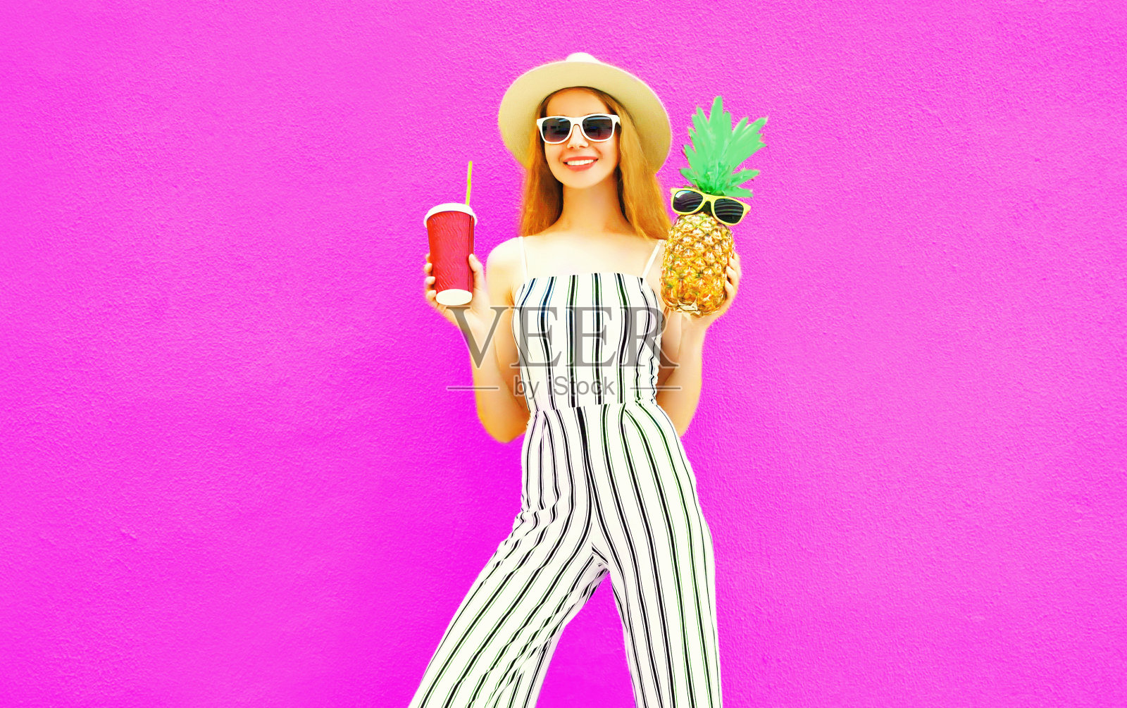快乐微笑的女子用菠萝，一杯果汁在夏天圆草帽，白色条纹连衣裤上彩色的粉红色墙壁背景照片摄影图片