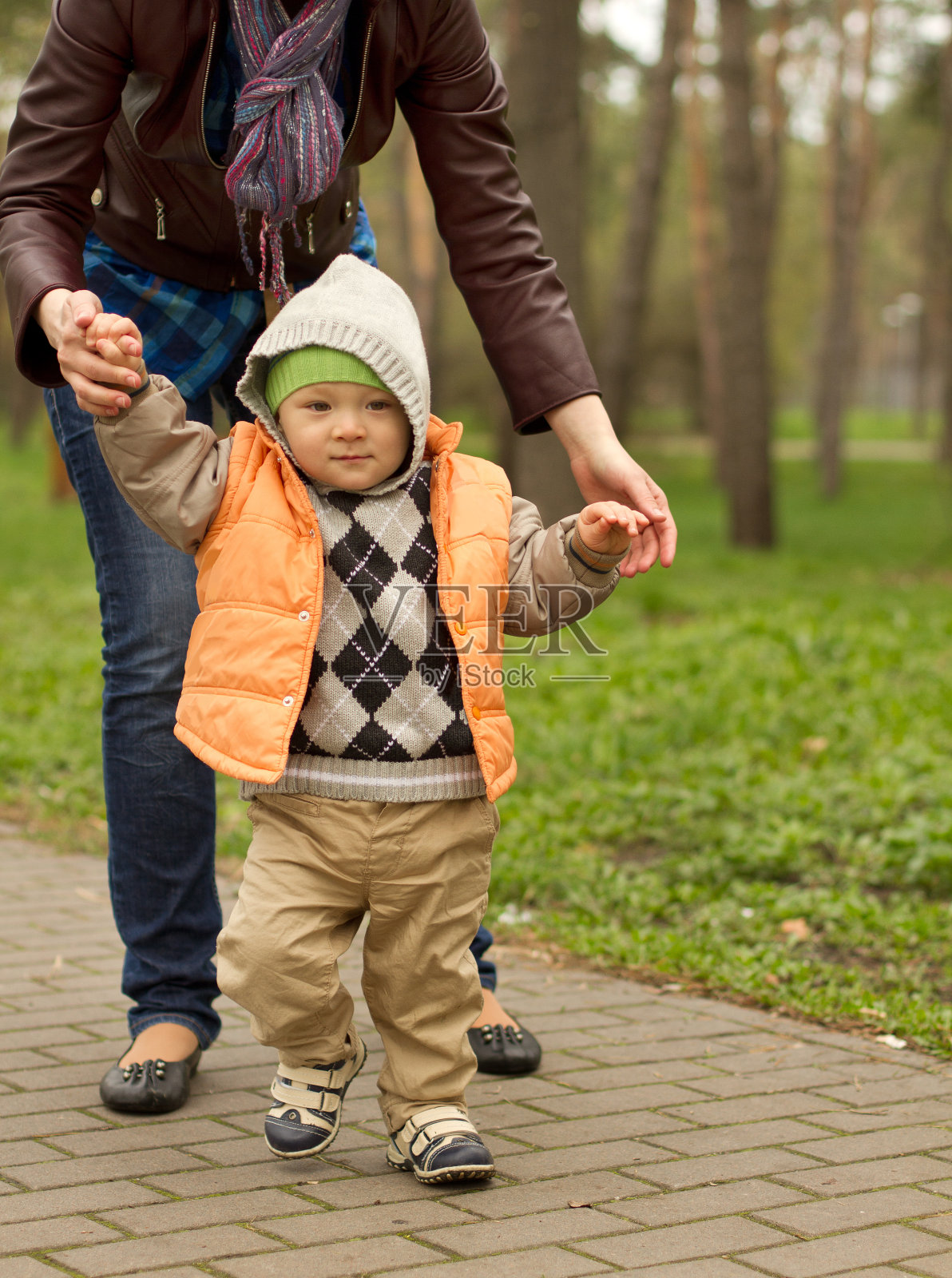 在公园里学走路的宝宝和他的妈妈在后面。可爱的小男孩在学走路。第一步是在母亲的帮助下照片摄影图片