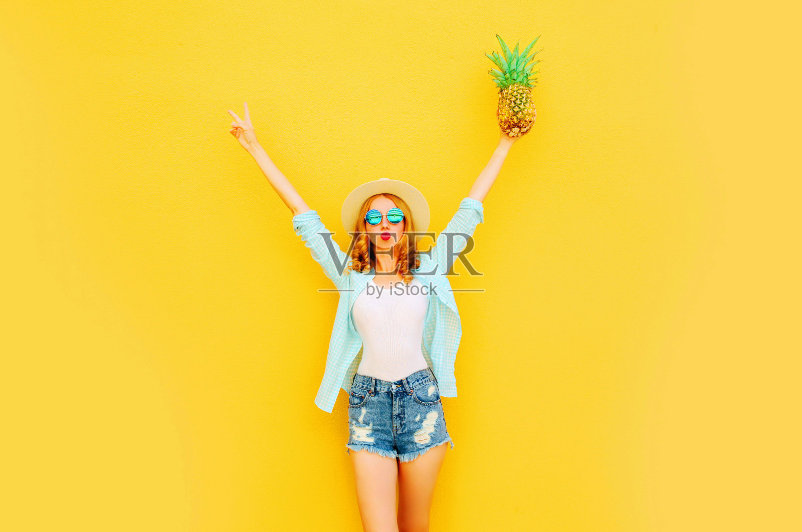 快乐的女人举起她的手与菠萝有乐趣的夏天草帽，太阳镜，短裤在彩色的黄色背景照片摄影图片