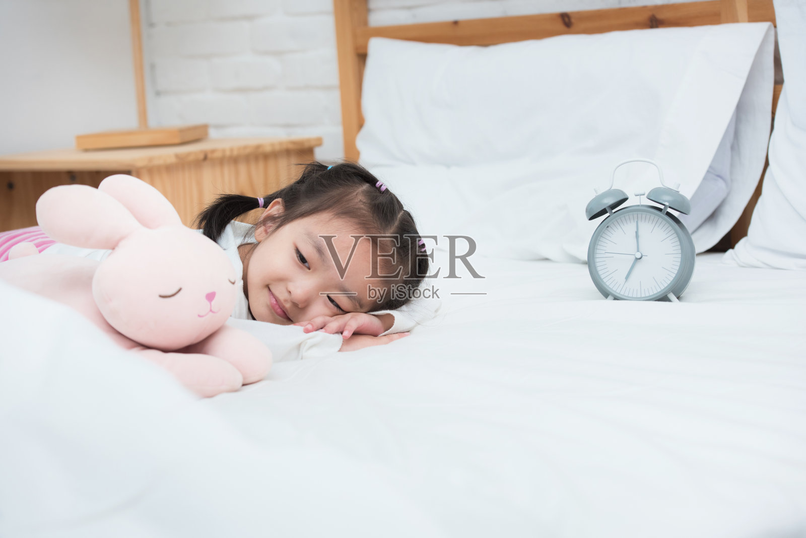 微笑的年轻亚洲女孩躺在床上的闹钟和娃娃照片摄影图片