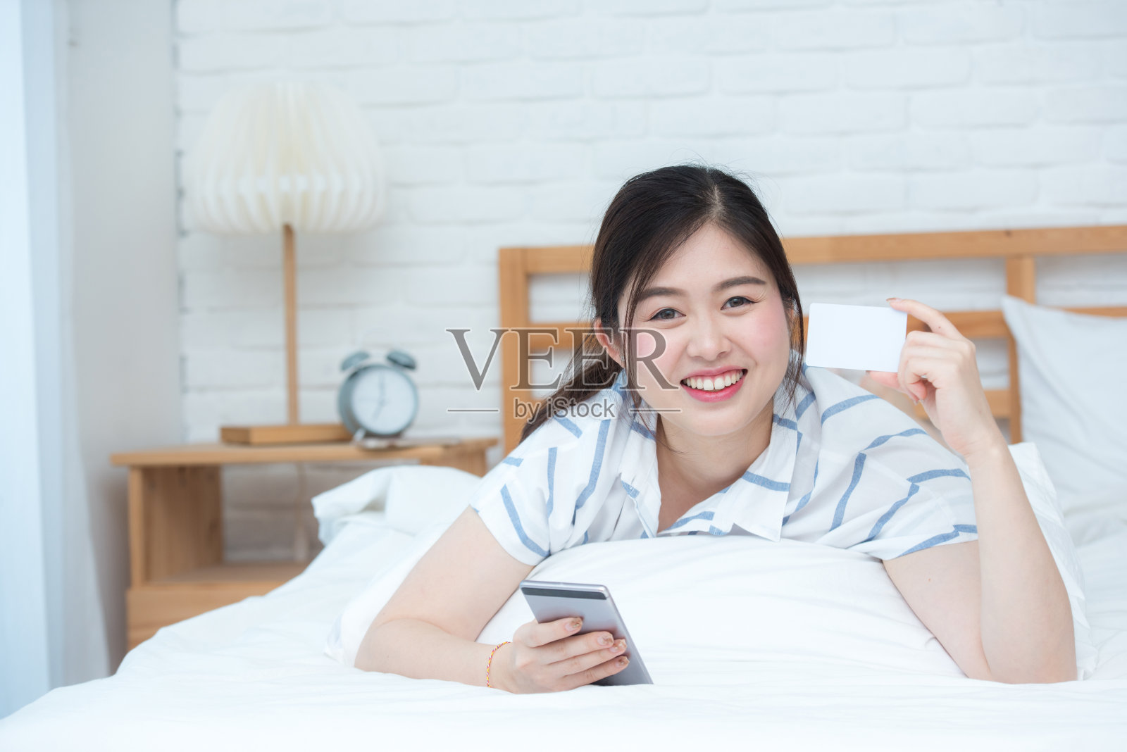 年轻美丽的亚洲女士在床上使用智能手机和信用卡。在线预订或在线商业概念。照片摄影图片