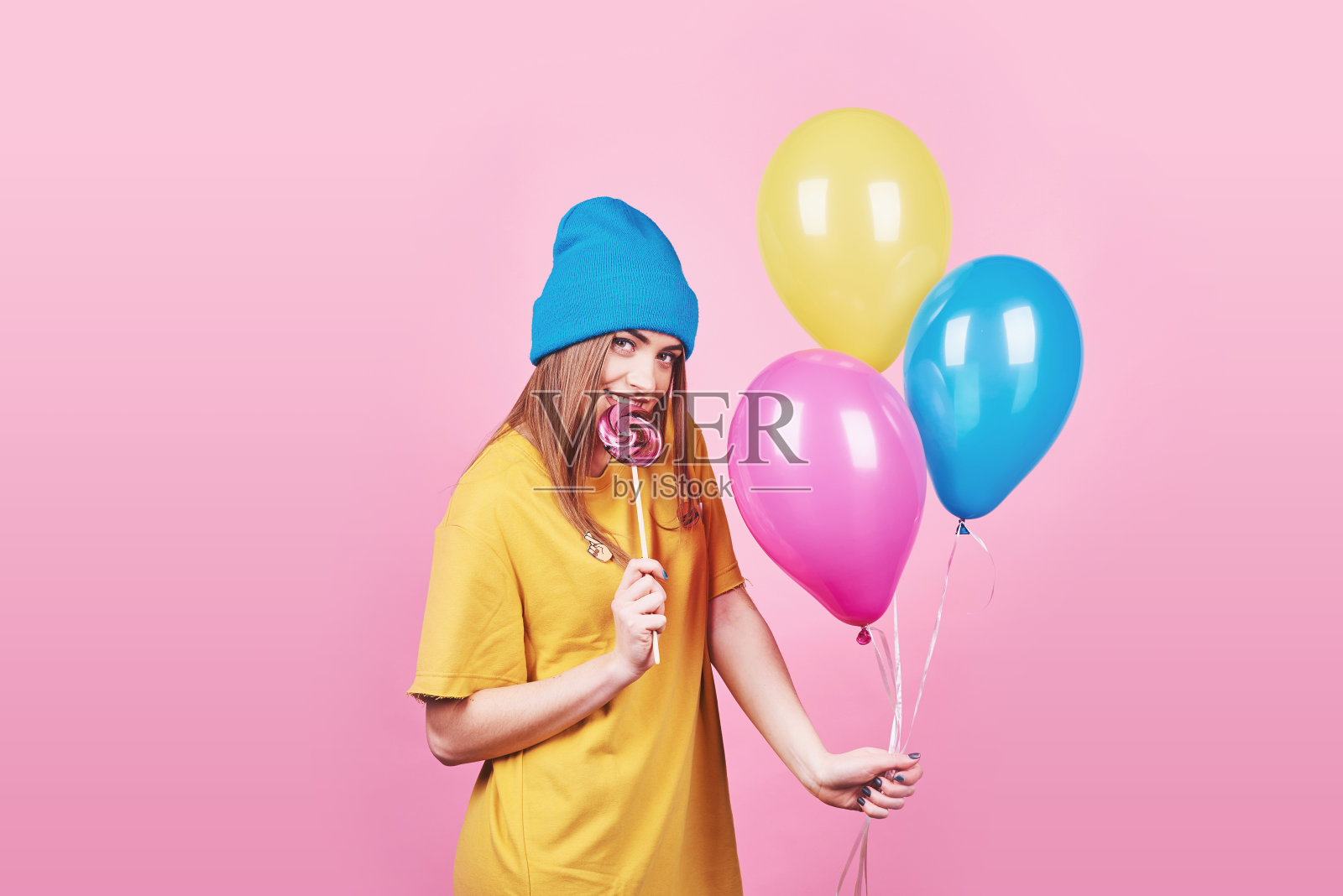 可爱有趣的女孩在蓝色的帽子肖像持有一个空气彩色气球和棒棒糖微笑在粉红色的背景。美丽的多文化白人女孩微笑快乐照片摄影图片