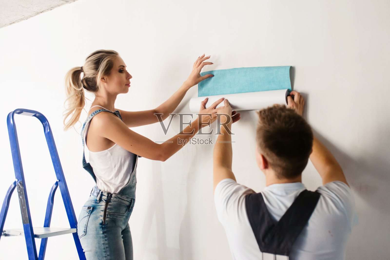 一对有趣的夫妇，一个女人和她的丈夫用一个金属梯子在墙上贴壁纸。家居装修和搬入新房的概念照片摄影图片