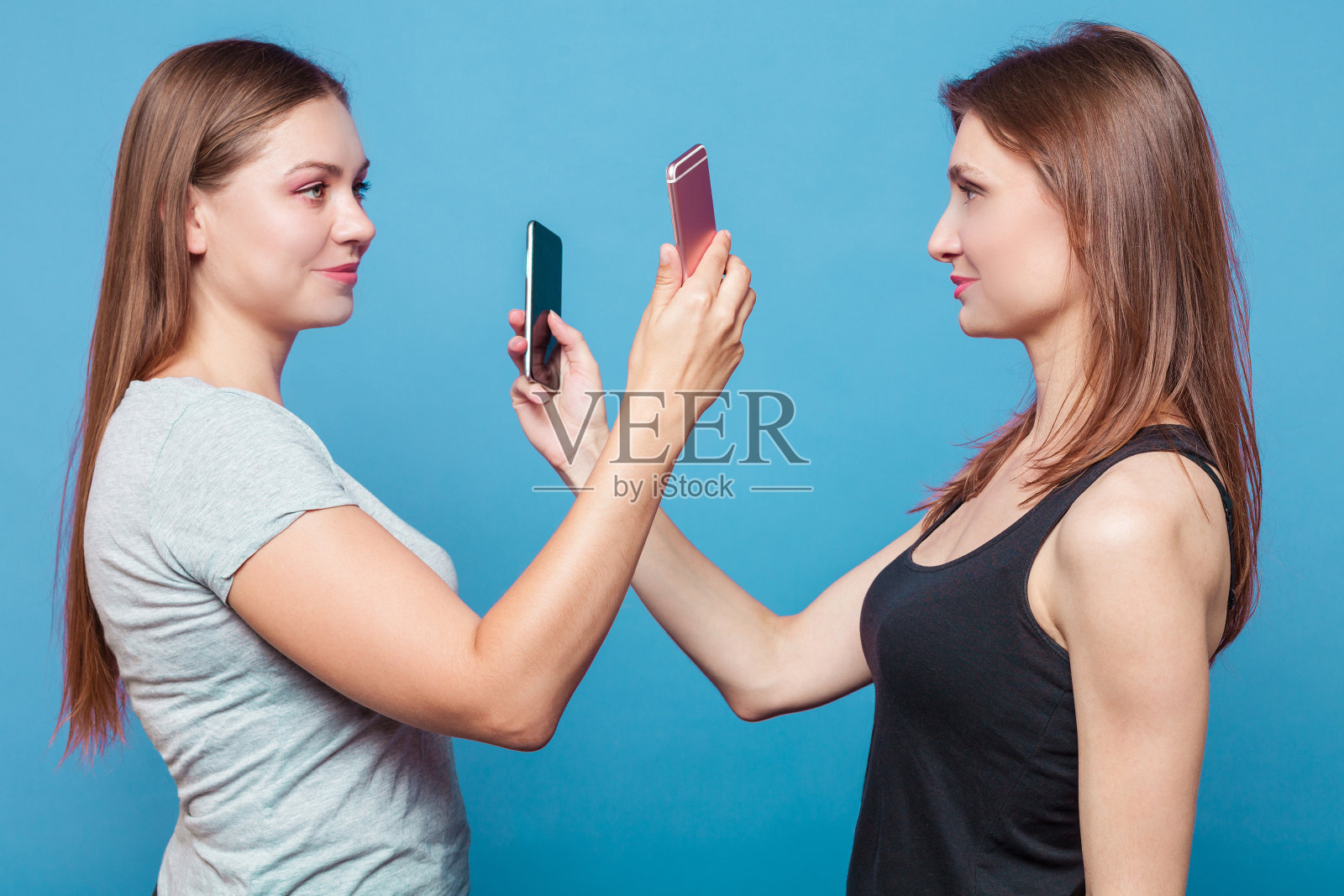 两个棕色头发的白人女孩面对面站着，互相拍手机照片。照片摄影图片
