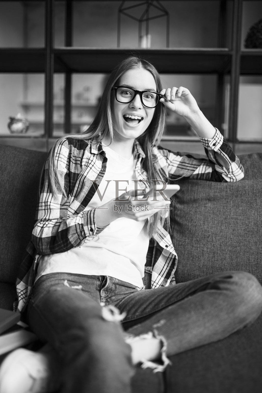 一个可爱的女孩戴着眼镜和手机在沙发上的肖像。黑色和白色照片摄影图片